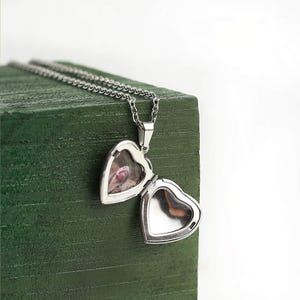 May include: A silver heart-shaped locket necklace, open to reveal a small photograph. The locket hangs from a delicate chain. The necklace is displayed on a textured, dark green surface, with a white background.