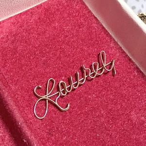 May include: A gold-colored wire word "Laurel" rests on a pink velvet surface inside a white jewelry box. The cursive script is delicate and elegant, perfect for a personalized gift or accessory.