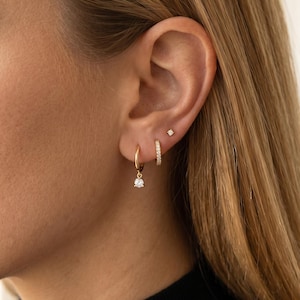 May include: Gold earrings on an ear. There is a small diamond stud, a small gold hoop with diamonds, and a gold hoop earring with a small diamond pendant. The earrings are gold in colour.