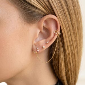May include: A collection of gold-toned earrings adorns an ear. The set includes a crescent moon stud, a star stud, a hoop earring with a pearl, and a chain connecting to a cuff earring. The cuff and hoop earrings feature small, sparkling embellishments.