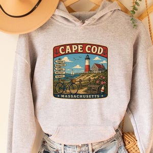 Op de afbeelding: Een lichtgrijze hoodie met een "Cape Cod Massachusetts"-afbeelding met een vuurtoren, fiets en wegwijzer. Een bruine hoed hangt aan de linkerkant. De hoodie wordt gecombineerd met een spijkerbroek. Een houten kleerhanger en een geweven tas zijn ook zichtbaar.