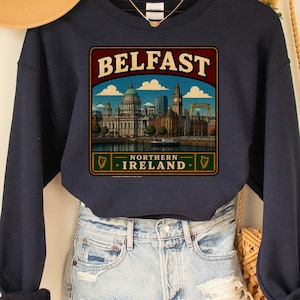 May include: A navy blue crewneck sweatshirt featuring a graphic print of the Belfast, Northern Ireland skyline. The design includes the text BELFAST and NORTHERN IRELAND with illustrations of city buildings, a harbor, and small harp icons.