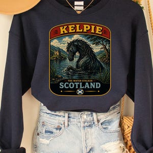 May include: A navy blue long-sleeve sweatshirt featuring a graphic print of a black horse in water with the text KELPIE THE WATER STALKER SCOTLAND. The sweatshirt is paired with light blue distressed denim shorts and a tan wide-brimmed hat.