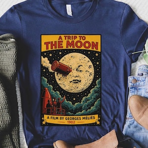 以下が含まれることがあります： ヴィンテージ映画ポスターのデザインが施されたネイビーブルーのTシャツ。「A Trip to the Moon」と書かれたポスターには、月とロケットのイラストが描かれています。「A Film by Georges Méliès 1902」というテキストも含まれています。