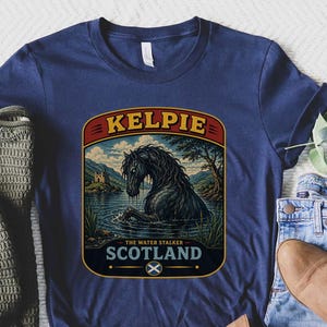May include: A navy blue t-shirt featuring a graphic of a black horse emerging from a lake with the text KELPIE THE WATER STALKER SCOTLAND and a small Scottish flag icon. The design is framed in a rounded rectangular border with red and gold accents.
