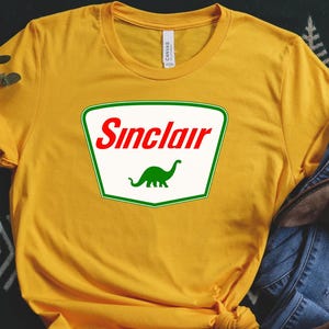 May include: Mustard yellow t-shirt with a Sinclair logo, featuring a green dinosaur. The logo is in a white and green rectangle with the word "Sinclair" in red. The shirt is styled with blue jeans and brown boots.