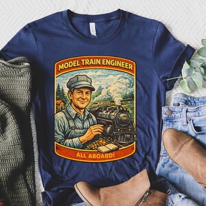 Puede incluir: Camiseta azul marino con una ilustración vintage de un ingeniero de trenes modelo. El gráfico incluye el texto "MODEL TRAIN ENGINEER" y "ALL ABOARD!". El diseño está enmarcado en un borde rojo y dorado. La camiseta se combina con vaqueros azules y botas marrones.
