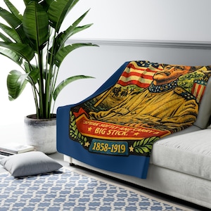 May include: A throw blanket featuring a vintage-style illustration of Theodore Roosevelt in military uniform against an American flag. The text reads, "SPEAK SOFTLY AND CARRY A BIG STICK." and "1858-1919" on a red and blue banner with laurel leaves.
