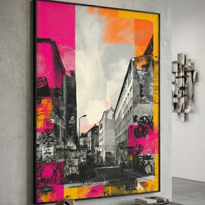 Puede incluir: Impresión artística de una escena callejera abstracta con marco negro. La obra presenta un collage colorido de edificios, grafitis y elementos callejeros en tonos rosa, naranja, amarillo y gris. La impresión se exhibe en una pared gris.