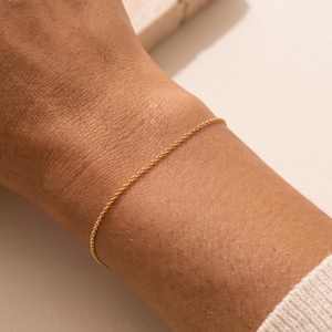 May include: A delicate gold chain bracelet featuring a fine, rounded link design that wraps around the wrist. The bracelet has a polished, reflective surface and a thin, minimalist profile, showcasing a continuous, uniform pattern of small, interconnected links.