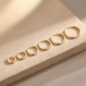 May include: A collection of six gold hoop earrings of varying sizes, arranged from smallest to largest. The earrings are displayed on a light beige surface, showcasing their polished, reflective finish. The hoops are round and have a simple, classic design.