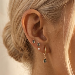 May include: A set of three gold earrings worn in one ear. The top earring is a small stud with a teardrop-shaped green stone. The middle earring is a stud with a teardrop green stone and a small clear stone. The bottom is a gold hoop with a teardrop green stone.