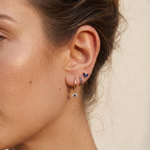 May include: Close-up of an ear featuring three earrings. A gold hoop with a blue eye charm, a gold hoop with blue stones, and a small blue butterfly stud. The earrings are set against a neutral background.