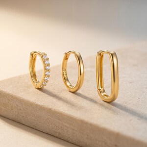May include: Three gold-toned hoop earrings arranged in a row. The first is a small circular hoop set with clear stones. The second is a plain circular hoop with S925 stamped on the side. The third is an elongated oval-shaped hoop earring.