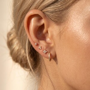 May include: A set of four gold-toned earrings worn in a single ear. The collection includes one small hoop earring, one four-stone flower-shaped stud, one three-stone triangular stud, and one single-stone stud, all featuring clear, sparkling stones.