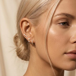 May include: An ear displaying three gold earring styles. The first is a trio of star studs, the second a floral design, and the third a small hoop. Each earring is decorated with tiny, sparkling gemstones. The earrings are a delicate and elegant accessory.