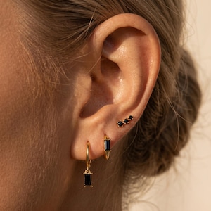 May include: A set of gold-toned earrings consisting of a small hoop with a dangling rectangular black stone, a small hoop with a fixed rectangular black stone, and a stud earring featuring three small round black stones in a row.