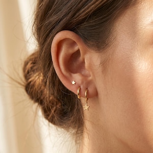 Könnte beinhalten: Ein Paar goldfarbene Ohrringe bestehend aus einem kleinen sternförmigen Stecker und einem dünnen Creolen-Ohrring mit einem hängenden Sternanhänger. Der Schmuck wird im Ohrläppchen getragen und zeigt eine polierte metallische Oberfläche.