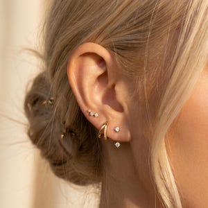 May include: A set of gold-toned earrings consisting of a small three-star stud, a double-hoop earring with a pavé-set band, and a single solitaire stud earring with a small dangling stone, all worn along the cartilage and earlobe.