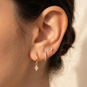 May include: A close-up of an ear featuring three gold earrings. There's a small stud, a small hoop with clear stones, and a gold hoop with a diamond-shaped charm hanging from it. The earrings are all gold in colour.
