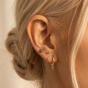 May include: A set of gold-toned earrings consisting of a four-leaf clover stud with green stones, a small round green stone stud, a plain gold hoop, and a gold hoop featuring a small dangling green stone charm, all worn in a single ear.