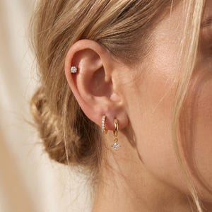 Könnte beinhalten: Ein goldfarbenes Ohr-Set bestehend aus einem runden Ohrstecker am oberen Knorpel und zwei kleinen Creolen am Ohrläppchen. Eine Creole ist schlicht goldfarben, während die andere einen kleinen, hängenden, quadratischen, klaren Edelsteinanhänger hat.