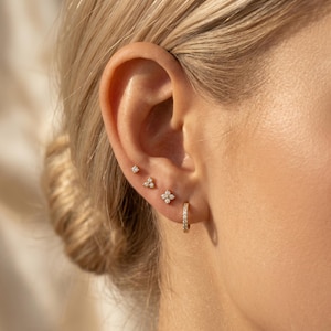 May include: Close-up of an ear adorned with multiple gold earrings. The ear has a small diamond stud, a cluster of three diamonds, a four-petal flower design, and a small hoop earring with embedded diamonds. The earrings are set against the backdrop of a person's skin and blonde hair.