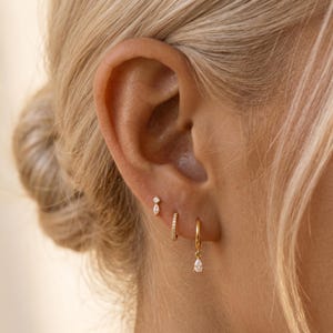 May include: A set of three gold earrings worn in a single ear. The top earring is a small stud with a marquise-cut stone. The middle earring is a thin gold hoop with pavé stones. The bottom earring is a gold hoop with a pear-shaped drop stone.