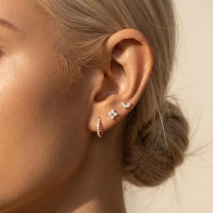 May include: Close-up of an ear with multiple gold earrings. A small hoop earring is in the lobe, a four-petal flower stud, and a marquise-shaped stud are visible. All earrings are embellished with small, sparkling stones.