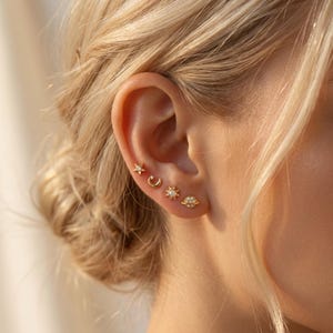 May include: Close-up of a person's ear adorned with four gold stud earrings. The earrings feature celestial designs: a star, a crescent moon, a sun, and a small spaceship. The earrings are small and delicate, with a shiny finish.