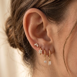 May include: A selection of gold earrings, including small stud earrings and hoop earrings. The studs feature a leaf design and a flower design. The hoops have dangling diamond-like accents. The earrings are worn on an ear.