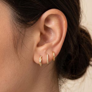 May include: Three gold hoop earrings are displayed on an ear. The first earring is a small twisted hoop with small clear stones. The second is a small hoop with clear stones. The third is a plain gold hoop.