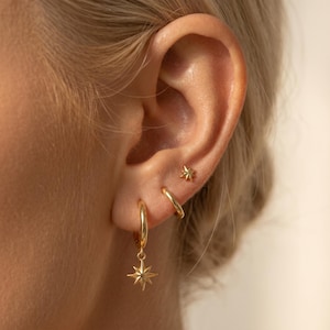 May include: A set of three gold-toned earrings consisting of one small star-shaped stud, one plain hoop, and one hoop featuring a dangling eight-pointed star charm. The jewellery is displayed in the earlobe and cartilage, showcasing a polished finish.