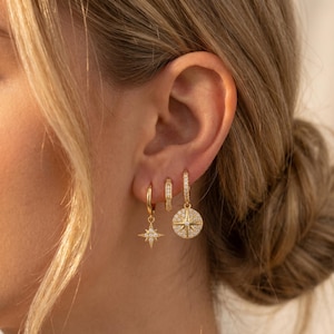 May include: Three gold-toned hoop earrings worn in a row. The first is a small hoop with a dangling starburst charm featuring clear stones. The second is a small hoop with a vertical row of clear stones. The third is a hoop with a round starburst charm.