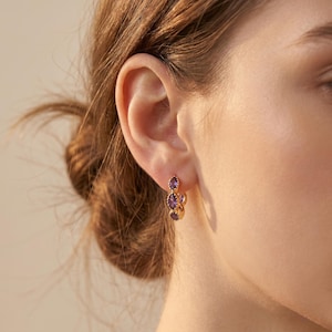 Könnte beinhalten: Goldfarbene Ohrringe mit drei lila Amethyst-Edelsteinen. Die Ohrringe sind im Tropfenstil gehalten, wobei jeder Edelstein in einen goldenen Rahmen gefasst ist. Die Ohrringe werden an einem Ohr getragen, wobei der Fokus auf dem Schmuck liegt.