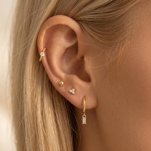 Könnte beinhalten: Nahaufnahme eines Ohrs mit mehreren goldenen Ohrringen. Das Ohr hat einen kleinen goldenen Kugelstecker, einen dreieckigen Ohrring mit drei kleinen Steinen, einen Creolen-Ohrring mit einem rechteckigen Anhänger und einen Helix-Ohrring mit einem einzelnen klaren Stein.