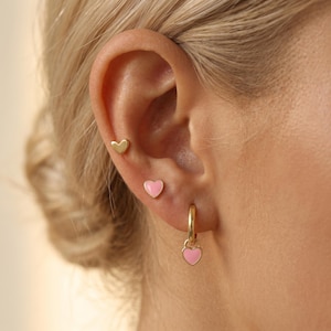 Könnte beinhalten: Ein Set aus drei goldfarbenen Ohrringen, bestehend aus einem kleinen schlichten goldenen Herzstecker, einem kleinen rosa Emaille-Herzstecker und einem goldenen Creolen-Ohrring mit einem baumelnden rosa Emaille-Herzanhänger, getragen an einem Ohrläppchen.