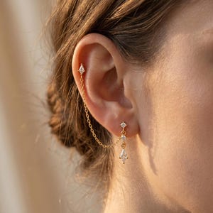 May include: Gold-coloured ear jewellery featuring a chain connecting two earrings. The upper earring is a small stud with a diamond-shaped gem. The lower earring has a dangling design with a diamond-shaped gem and two smaller gems.