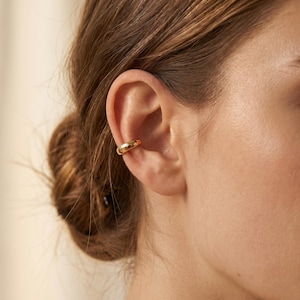 May include: A gold-colored ear cuff is worn on the ear. The ear cuff is a thick, rounded band that wraps around the ear cartilage. The skin tone is fair, and the hair is pulled back in a bun.
