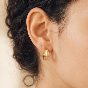 Peut inclure: Une petite boucle d'oreille créole en or au design triple anneau, composée de trois fils d'or arrondis, parallèles et polis, courbés pour former une forme circulaire continue. La boucle est fixée au lobe, affichant une finition métallique brillante.