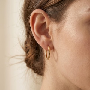 May include: A close-up shot of a person's ear with a gold hoop earring. The earring is a simple, circular design, and the gold color is bright and shiny. The earring is approximately 1 inch in diameter.