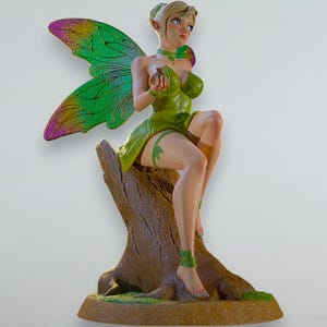 Puede incluir: Figura de hada con cabello rubio, vestido verde en forma de hoja y alas coloridas. El hada está sentada sobre un tocón de árbol marrón. Las alas son verdes con detalles amarillos y morados. El hada sostiene un objeto pequeño.