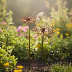 Puede incluir: Dos estacas de jardín de metal oxidado con forma de flor, con un diseño detallado de pétalos y un centro esférico. Las estacas están plantadas en el suelo de un jardín lleno de flores coloridas y vegetación, creando una estética rústica y natural.