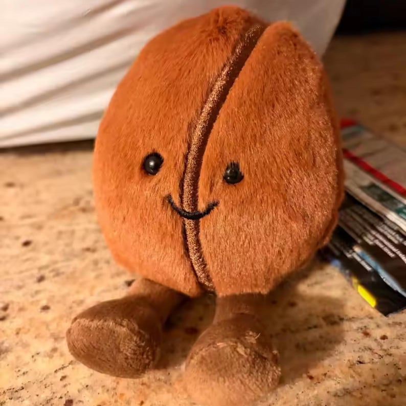 May include: A soft, brown coffee bean plush toy with a smiling face and small feet. The bean is split down the middle and has a fuzzy texture. The toy is designed to resemble a cheerful coffee bean.