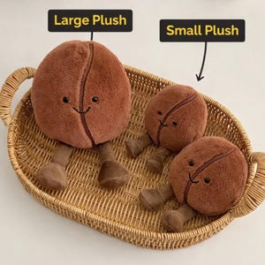 May include: Three plush coffee bean toys in a woven basket. The toys are brown with smiling faces and small legs. The text "Large Plush" and "Small Plush" are above the toys.