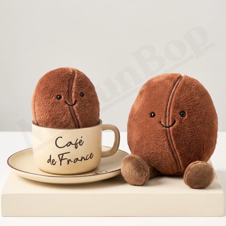 May include: Two plush coffee bean toys with smiling faces. One bean sits in a beige mug and saucer set with "Caf&eacute; de France" printed on the mug. The other bean sits upright. The beans are brown.