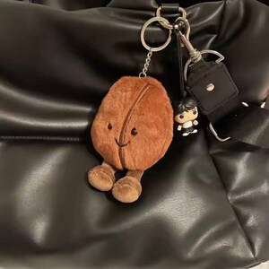 May include: A brown plush coffee bean keyring with a smiling face and small legs. A small figurine and a silver keyring and clasp are attached. The keyring hangs against a black leather bag.