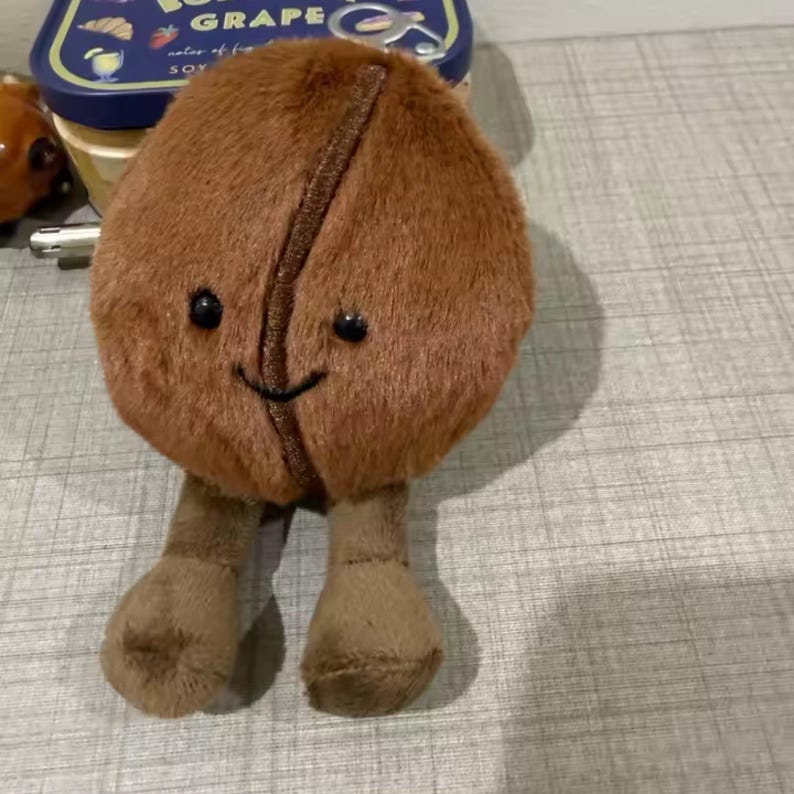 May include: A brown plush coffee bean character with a smiling face and small black eyes. The bean has two small brown legs. The character is sitting on a grey surface.