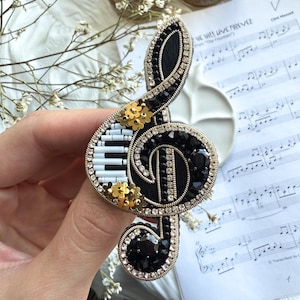 Könnte beinhalten: Schwarze und goldene Notenschlüssel-Brosche mit Klaviertasten-Design. Die Brosche ist mit schwarzen und klaren Kristallen und kleinen goldenen Blumenakzenten verziert. Die Brosche befindet sich auf einer weißen Oberfläche mit Noten im Hintergrund.