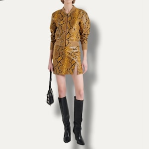 Peut inclure: Un blouson bomber et une mini-jupe assortie à motif peau de serpent jaune et noir, avec une fermeture éclair frontale et une fente latérale. L'ensemble est complété par des bottes noires arrivant au genou et un petit sac à main noir.